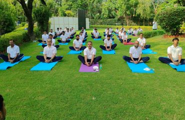 RCI officials practicing yoga outdoors on mats in butterfly pose during Yoga Day celebration.