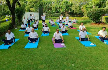 RCI officials seated on yoga mats in an outdoor garden practicing yoga during Yoga Day celebration.