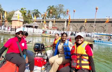 सणाच्या वेळी मंदिराचे सजलेले घाट आणि पार्श्वभूमीत भाविक दिसत असताना सिद्धेश्वर मंदिर तलावावरील बचाव नौकेवर जीवरक्षक जॅकेट घातलेल्या आपदा सख्या आणि पोलीस कर्मचारी हसत असताना