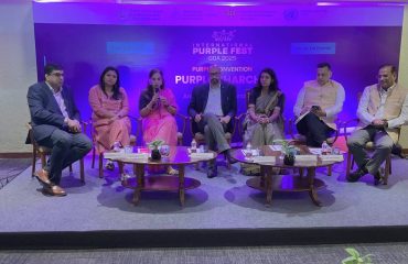 A panel of seven speakers is seated on a stage during the International Purple Fest 2025 at Taj Vivanta. They are engaged in a discussion under the banner “Purple Charcha,” organized by the State Commission for Persons with Disabilities – Goa, Department of Empowerment of Persons with Disabilities, and the United Nations. The panel includes men and women in formal attire, with one woman speaking into a microphone. The backdrop features the event name, logos of organizing bodies, and the date “10th Oct 2025.” Small tables with water bottles and plants are placed in front of the panelists.
